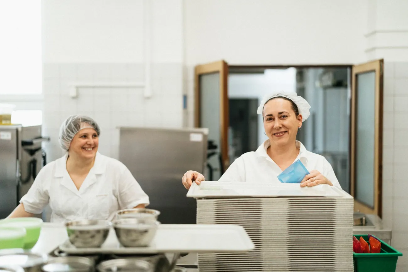 Two canteen staff in white uniforms and hairnets at the serving counter of an industrial kitchen, with stacked meal trays